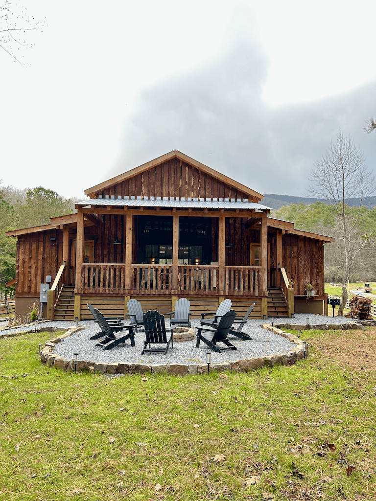 Front facade of The Barn at Haynes Hill — board-and-batten siding, covered porch, fire pit ring with adirondack chairs in the foreground, mountains in the misty distance