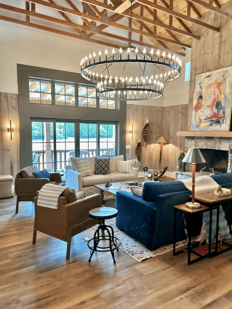 The great room at The Barn at Haynes Hill — a chandelier of forged iron, exposed timber trusses, layered seating around a stone hearth, and floor-to-ceiling windows