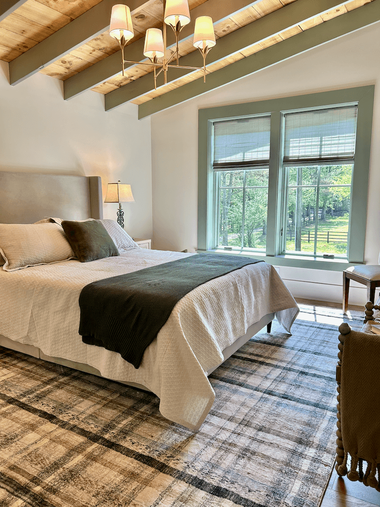 Master bedroom at The Barn — king bed dressed in olive and cream linens, large windows facing the pasture, exposed beams overhead