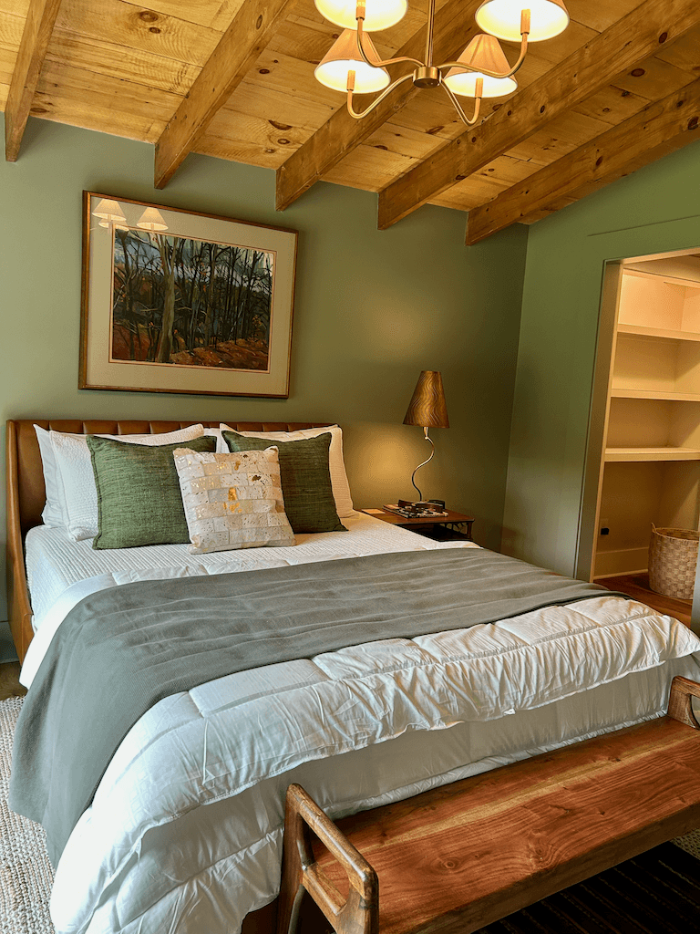 Sage guest room — queen bed beneath a framed landscape, plank ceiling, built-in shelving, and a small bench at the foot of the bed