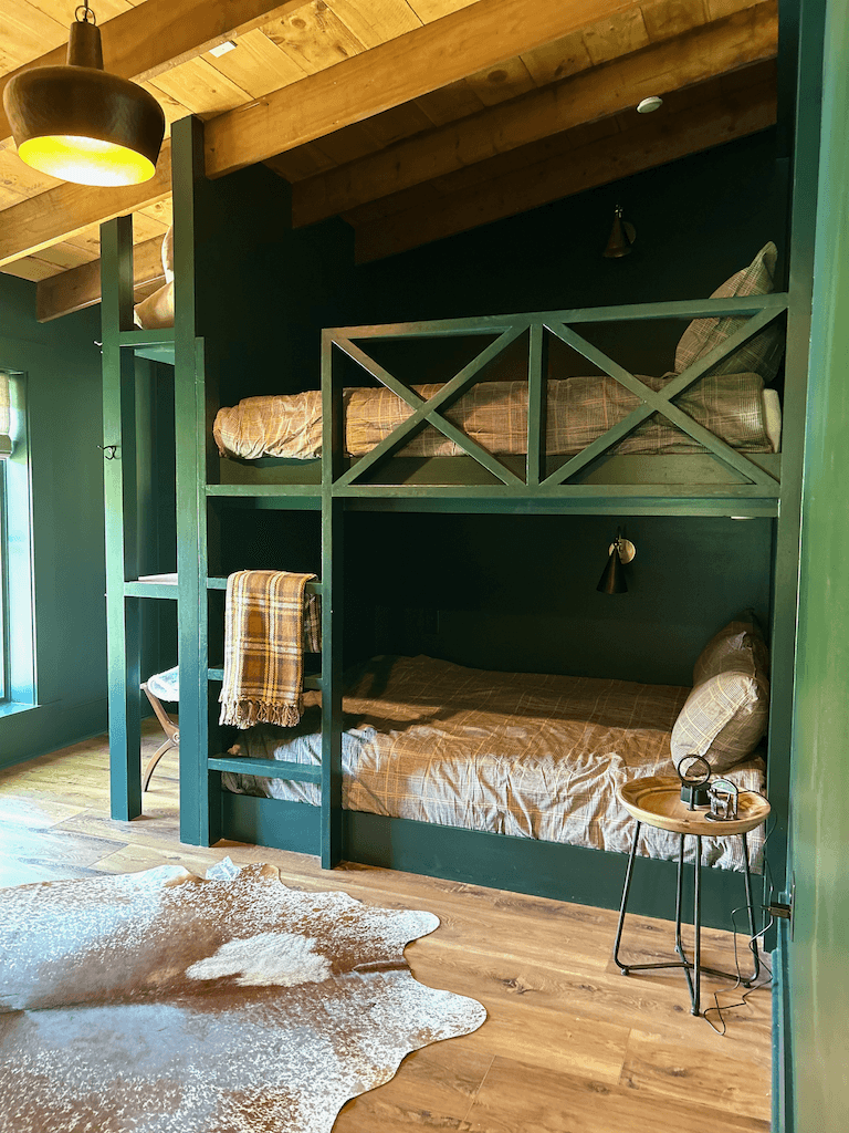 The bunk room — built-in twin bunks painted forest green with white-cross side rails, leather chair, cowhide rug on wide-plank floors