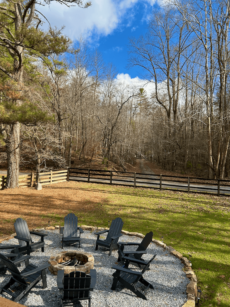 The fire pit circle — adirondack chairs arranged around a stone fire pit on a gravel surround, split-rail fence and forest beyond