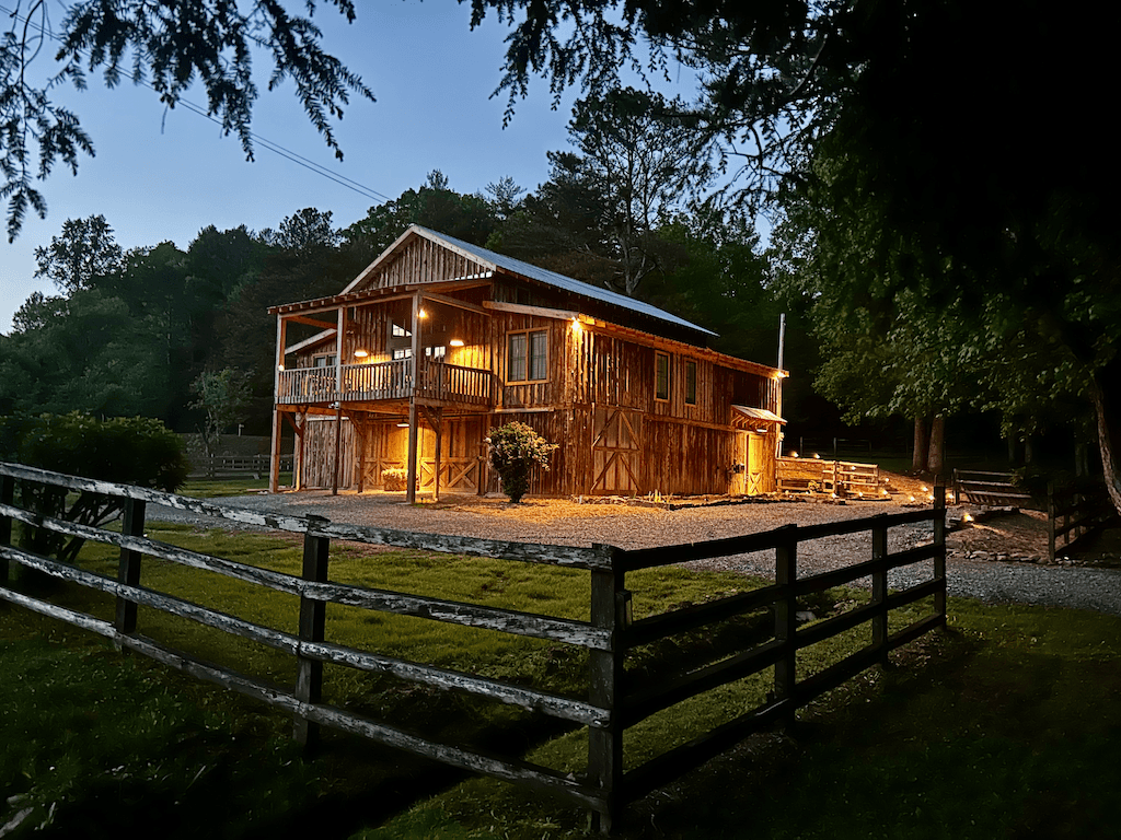 The Barn at Haynes Hill at dusk — warm porch and interior lights glowing through the open ground floor as the day ends